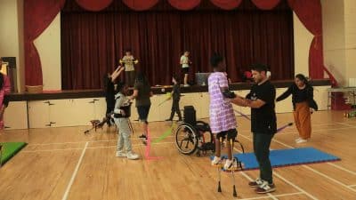 Students stiltwalking and working with poi and flowersticks in a community hall with a red curtain in the background.