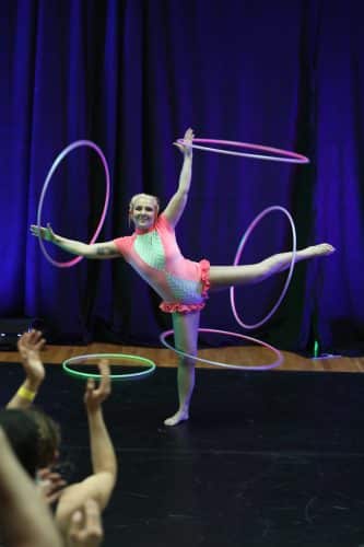 Hoopixie execuses a "Box splits". Standing on one leg with a hula hoop spinning on her knee, one leg extended to the side with another hoop on her foot, and arms above her head and one to he side also spinning hula hoops. wearing a pink, white and green leotard with ruffles along her thighs.