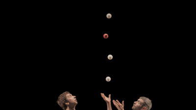 Two male performers juggling balls on a rolling platform against a black background, part of a contemporary circus act