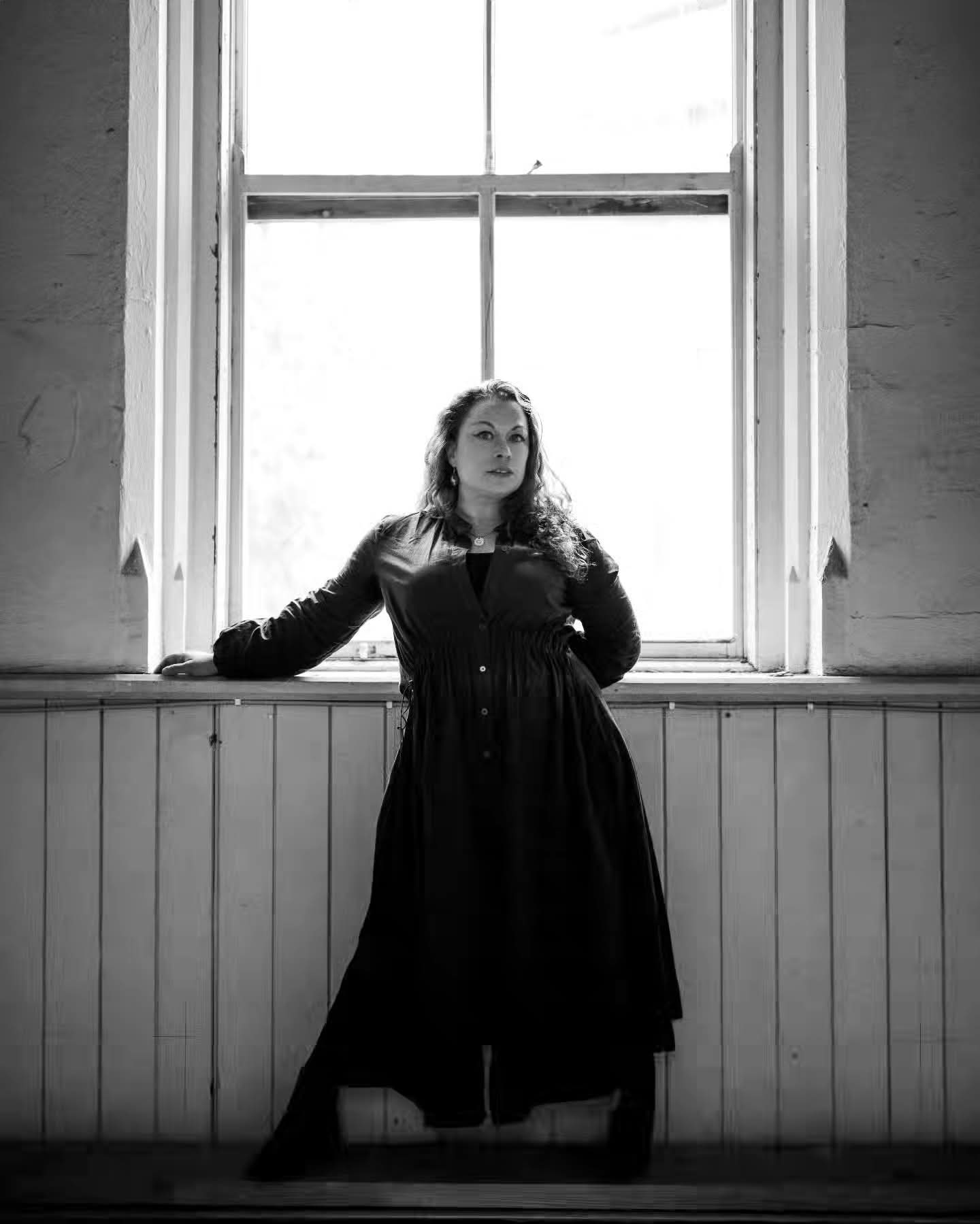 A black and white photo of a woman in a room standing in front of a window.