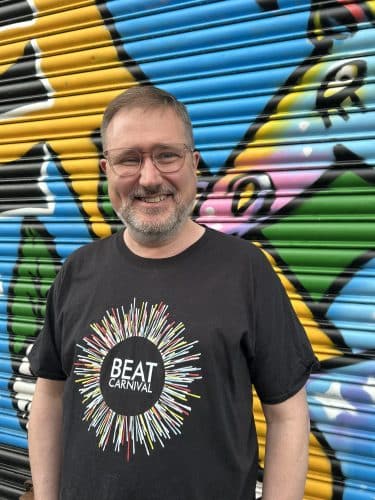 A man wearing glasses smiles for the camera. He is wearing a black t-shirt which has the Beat Carnival circular logo on it. He stands in front of a multi-coloured exterior shutter.