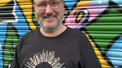 A man wearing glasses smiles for the camera. He is wearing a black t-shirt which has the Beat Carnival circular logo on it. He stands in front of a multi-coloured exterior shutter.